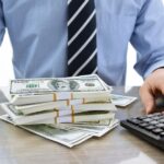 man with hands on table with pile of cash in front of him and a calculator