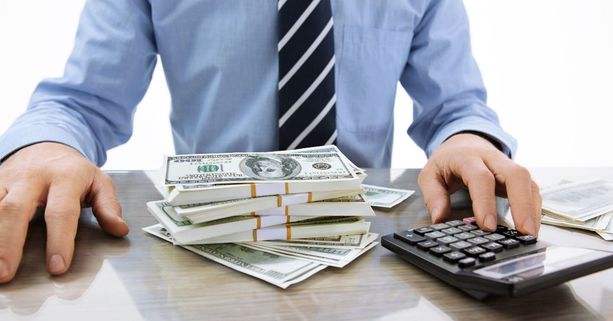 man with hands on table with pile of cash in front of him and a calculator