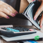 A person with pink painted fingernails is looking inside an empty wallet while a calculator lies in front of her. This implies she is struggling with money.