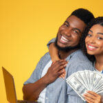 A man and woman are standing together, smiling, hands touching. He is holding a laptop computer and she is holding cash.